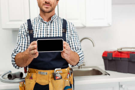 Cropped view of plumber holding smartphone with blank screen in kitchenの写真素材