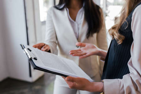 cropped view of businesswoman pointing with hand at clipboard near coworker in office