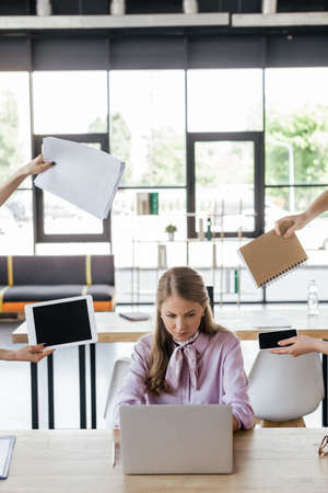 stressed businesswoman using laptop near coworkers holding gadgets with blank screen, notebook and papersの写真素材