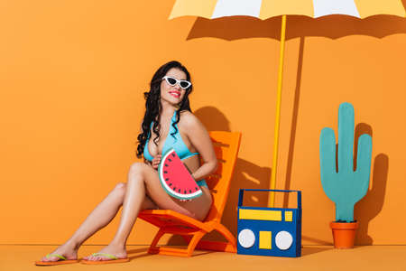 cheerful girl in sunglasses and swimsuit sitting on deck chair near boombox, cactus and umbrella...