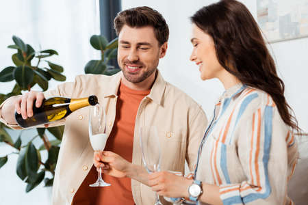 Selective focus of smiling man pouring champagne in glass near beautiful girlfriend at home
