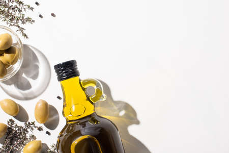 top view of olive oil in bottle near green olives in bowl, herb and black pepper on white background