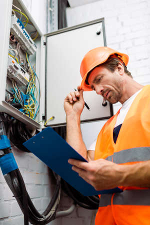 Selective focus of handsome electrician in hardhat holding pen and looking at clipboard near electric panelの写真素材