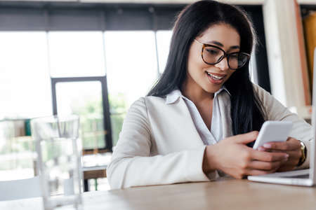 selective focus of happy businesswoman using smartphone near laptopの写真素材
