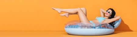 panoramic crop of barefoot woman in sunglasses and swimwear lying on inflatable ring on orangeの写真素材