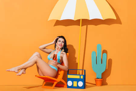 smiling woman in swimwear sitting on deck chair near paper boombox, cactus and umbrella while holding cocktail on orangeの写真素材