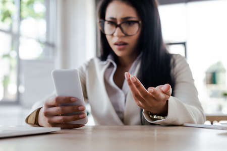 selective focus of displeased businesswoman using smartphone and gesturing in officeの写真素材
