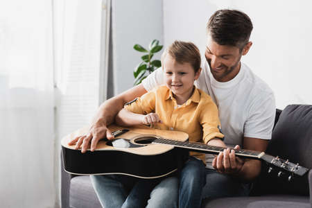 smiling boy sitting of fathers knees and learning how to play acoustic guitarの写真素材