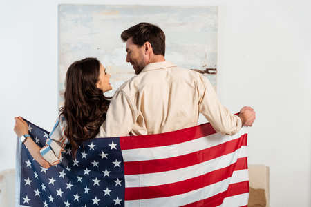 Back view of young couple smiling at each other while holding american flag at homeの写真素材