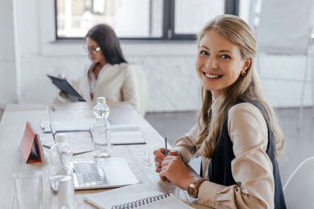 selective focus of happy businesswoman looking at camera near laptop and coworker in office