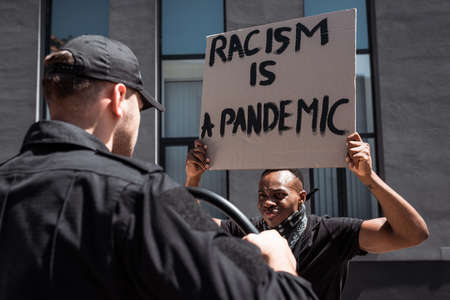 selective focus of african american man holding placard with racism is a pandemic lettering near...