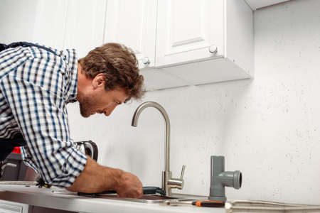 Side view of plumber holding pipe wrench while repairing kitchen faucet near tools on worktop