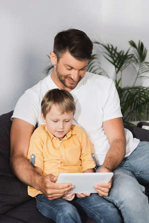 happy father with attentive son sitting on sofa and using laptop togetherの写真素材