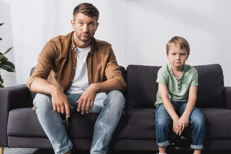 KYIV, UKRAINE - JUNE 9, 2020: upset father and son looking at camera while sitting on sofa with joysticksの写真素材
