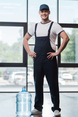 cheerful delivery man in uniform standing with hands on hips near bottled waterの写真素材