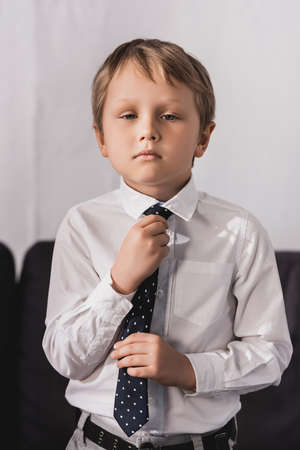 serious, cute boy in white shirt putting tie on and looking at cameraの写真素材