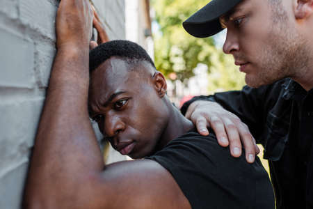 selective focus of policeman in cap detaining african american man on street, racism concept