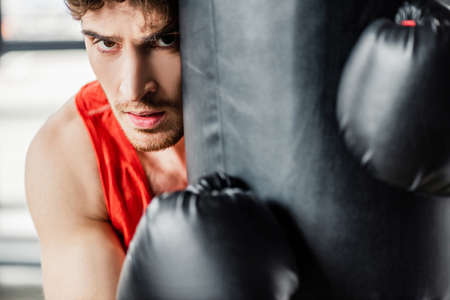 selective focus of tired man in sportswear and black boxing gloves touching punching bag in gym