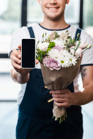 cropped view of happy delivery man holding bouquet of blooming flowers and smartphone with blank screenの写真素材