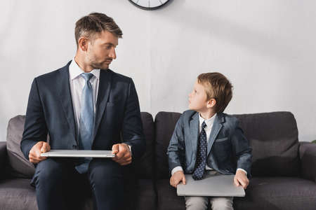 serious businessman and his son in formal wear sitting with laptops on sofa at homeの写真素材