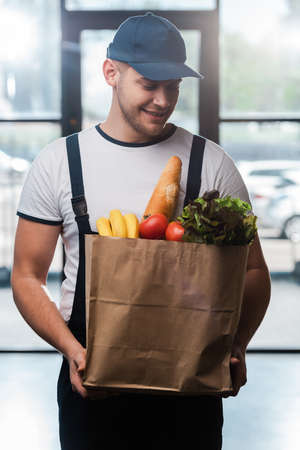 happy delivery man in cap looking at paper bag with groceriesの写真素材