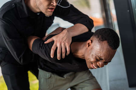 angry police officer detaining african american man suffering from pain, racism concept