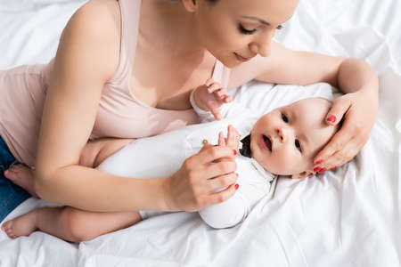 overhead view of attractive mother touching adorable infant son in baby romper lying on bed
