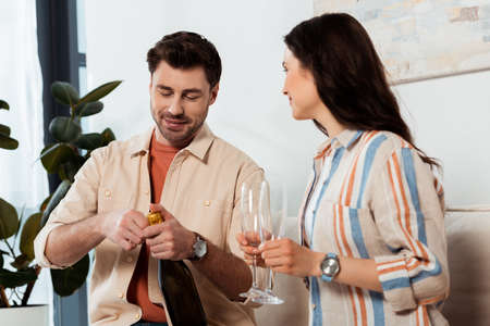 Handsome man opening bottle of champagne near woman holding glasses at home