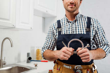 Cropped view of plumber in workwear and tool belt holding metal pipe in kitchenの写真素材
