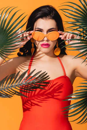 young and stylish woman touching sunglasses near green palm leaves isolated on orange