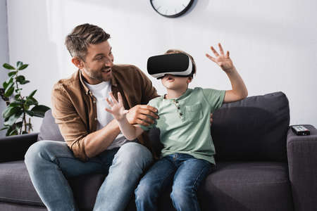 smiling father touching surprised son using vr headset and gesturing whiles sitting on sofaの写真素材