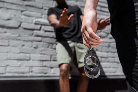 cropped view of police officer holding handcuffs while detaining african american man gesturing near brick wall, racism conceptの写真素材