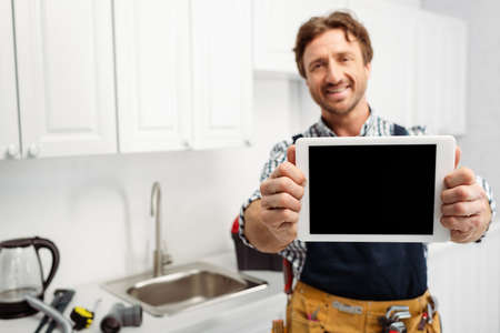 Selective focus of smiling plumber showing digital tablet with blank screen in kitchenの写真素材