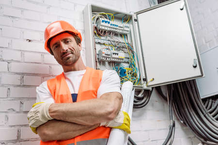 Handsome workman in helmet and safety vest holding blueprint and looking at camera near electric...