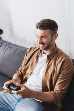 KYIV, UKRAINE - JUNE 9, 2020: happy, handsome man sitting on sofa and playing video game with...