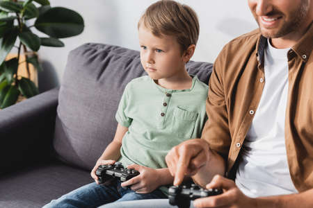 KYIV, UKRAINE - JUNE 9, 2020: cropped view of smiling man and attenive son playing video game wih joysticksの写真素材