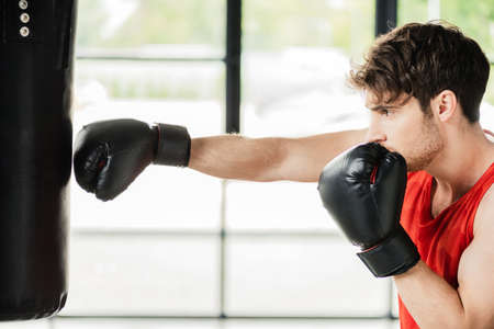 side view of athletic sportsman in boxing gloves working out with punching bagの写真素材