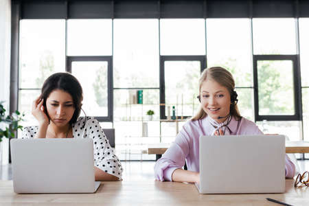 happy operator in headset smiling while looking at laptop near coworker