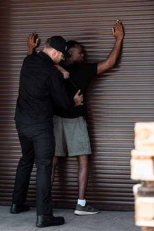 selective focus of police officer in uniform and cap holding truncheon while detaining african...