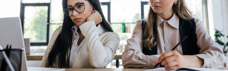 panoramic concept of bored businesswoman looking at laptop near coworker in officeの写真素材