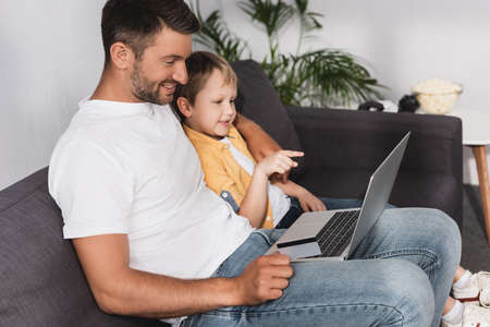 smiling boy pointing with finger at laptop while sitting near smiling father