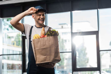 happy delivery man touching cap and holding paper bag with groceriesの写真素材