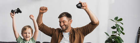 horizontal image of excited father and son holding joysticks and showing winner gestures