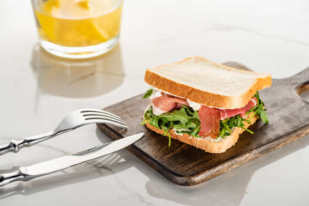 fresh green sandwich with arugula and prosciutto on wooden cutting board with cutlery on white marble surfaceの写真素材