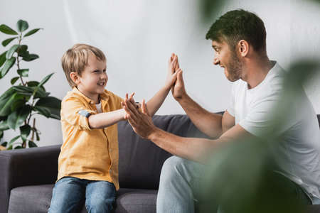 selective focus of cheerful father and son giving high five while sitting on sofa at homeの写真素材