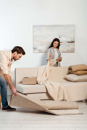 Selective focus of woman holding plaid near couch while boyfriend turning off carpet in living roomの写真素材