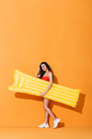 happy woman in swimsuit and sneakers holding inflatable mattress and touching hair on orangeの写真素材