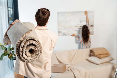 Back view of man holding carpet while girlfriend taking painting from wall during movingの写真素材