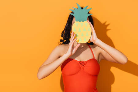 woman in swimsuit covering face with paper cut pineapple on orangeの写真素材