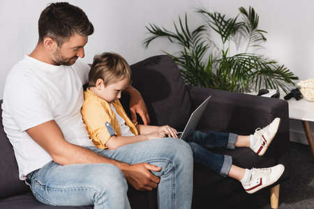 adorable boy using laptop while sitting on sofa together with fatherの写真素材